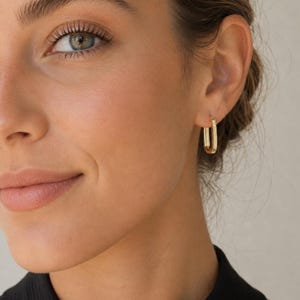 May include: A gold-toned rectangular hoop earring with a textured, ribbed surface along the outer edge. The earring features a hinged closure and is worn in the earlobe, showcasing a polished metallic finish and a structured, elongated geometric shape.