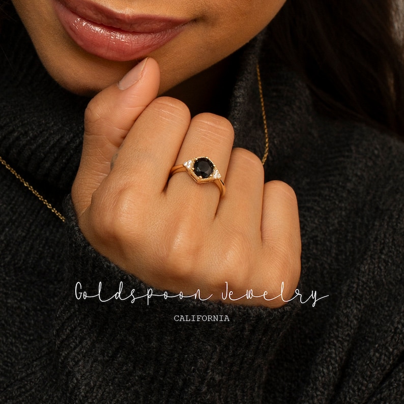 以下が含まれることがあります： 黒い宝石が小さな白い宝石で囲まれた金の指輪。指輪は金のバンドにセットされています。指輪は指にはめられています。背景には「Goldspoon Jewelry California」の文字が見えます。