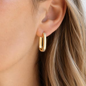 Könnte beinhalten: Ein goldfarbener Creolen-Ohrring in länglicher, rechteckiger Form mit einer texturierten, gerippten Oberfläche. Der Ohrring hat ein poliertes Finish, ist am Ohrläppchen befestigt und zeigt eine vertikale Ausrichtung mit abgerundeten Ecken.