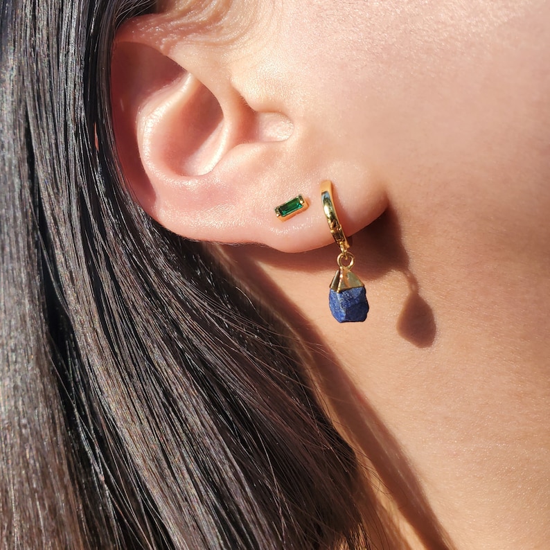 May include: Gold hoop earrings with a dangling blue gemstone. A smaller green gemstone earring is also visible.