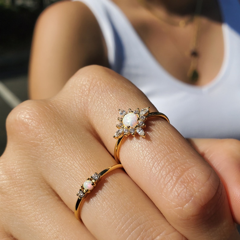 May include: Two gold rings with opalescent stones and small clear crystals. One ring features a central opal surrounded by crystals in a floral design. The other ring has a row of opals and crystals. Jewelry.