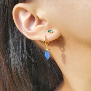 May include: Gold hoop earrings with a blue crystal pendant. The earrings are worn in the left ear. The crystal pendant is shaped like a teardrop.