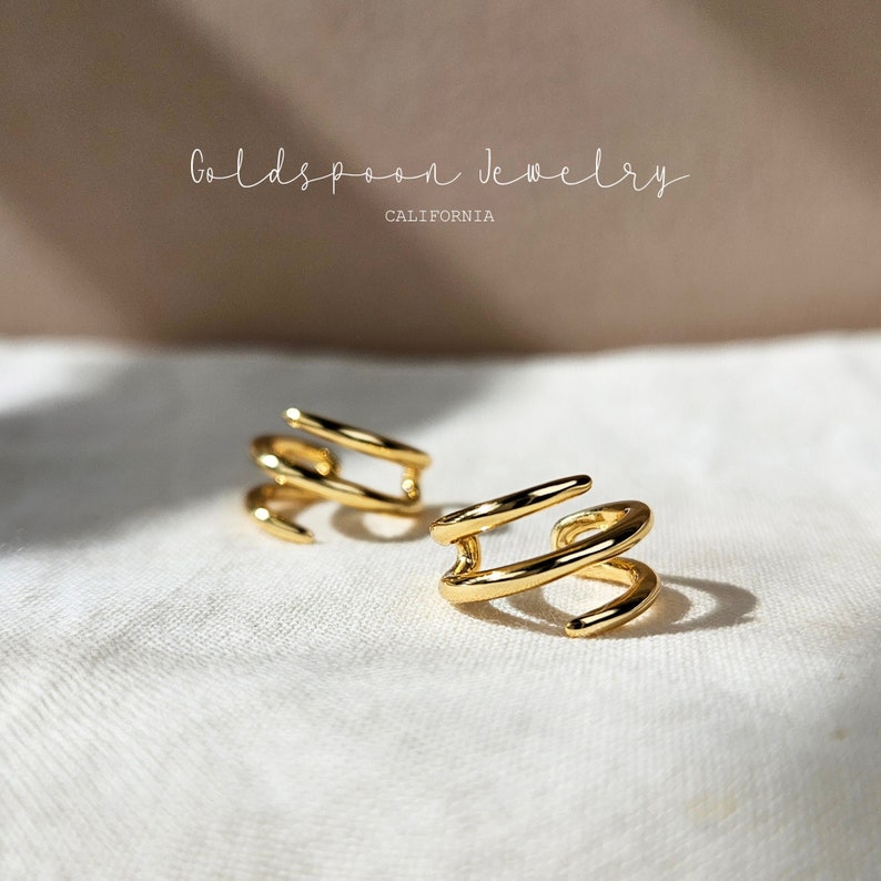 May include: Two gold ear cuffs with a simple, modern design. The cuffs are made of a single, continuous piece of metal that is twisted and shaped to create a unique and stylish look.