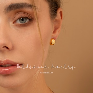 May include: A close-up of a woman's ear wearing a pair of gold hoop earrings. The earrings are simple and minimalist, with a smooth, rounded shape. The text "Goldspoon Jewelry" and "Minimalist" is visible below the earrings.