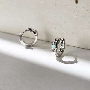 May include: A pair of small silver hoop earrings featuring a polished metallic finish and a single round, iridescent white opal stone set into the front of each hoop. The earrings have a hinged closure mechanism and a smooth, rounded design.