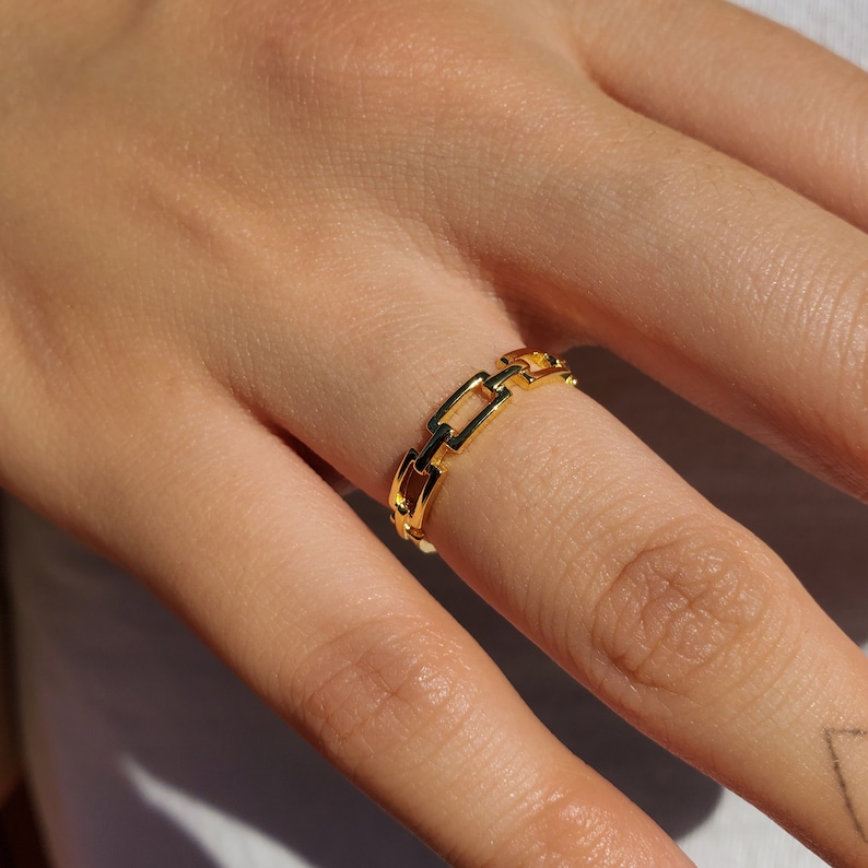 May include: A gold chain link ring. The ring features a series of rectangular links, forming a continuous chain pattern. The ring is worn on a finger, and the gold colour reflects light, giving it a shiny appearance. The ring is a piece of jewellery.