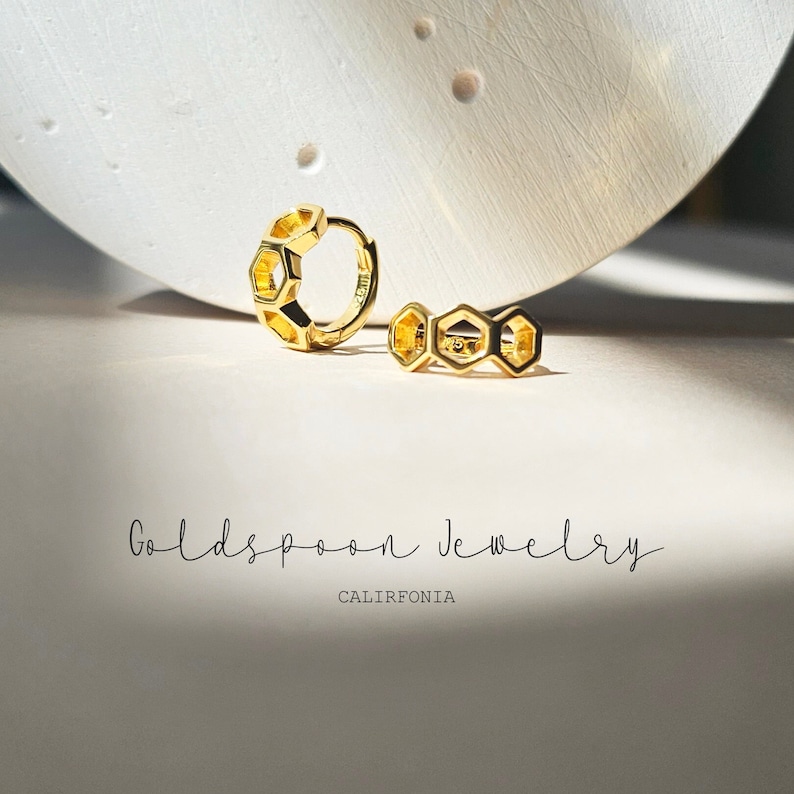 May include: A pair of gold hoop earrings with a honeycomb design. The earrings are on a white surface with the text "Goldspoon Jewelry California" below them.