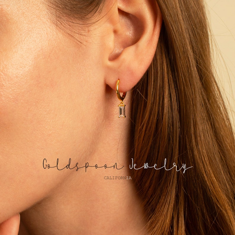 Puede incluir: Aretes de aro dorados con una piedra transparente rectangular colgante. Los aretes son usados por una persona con cabello largo y casta&ntilde;o. El texto "Goldspoon Jewelry California" es visible en la parte inferior de la imagen.