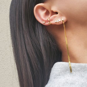 May include: Gold earrings with a long chain dangling from one ear. The earrings feature small, clear stones.