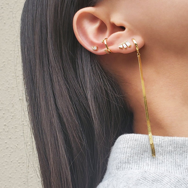 May include: Gold earrings with a long chain dangling from one ear. The earrings feature small, clear stones.