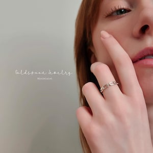 May include: A silver chain link ring is worn on a finger. The ring is in focus, with the wearer's face and hand partially visible. The text "Goldspoon Jewelry" and "Minimalist" are in the background.