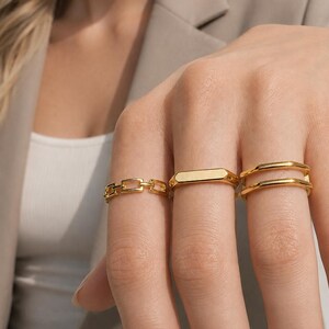 May include: Three gold-toned rings worn on fingers. One ring features a rectangular chain-link design, the second is a smooth flat-topped band, and the third is a double-layered band with a polished finish and a consistent metallic gold color.