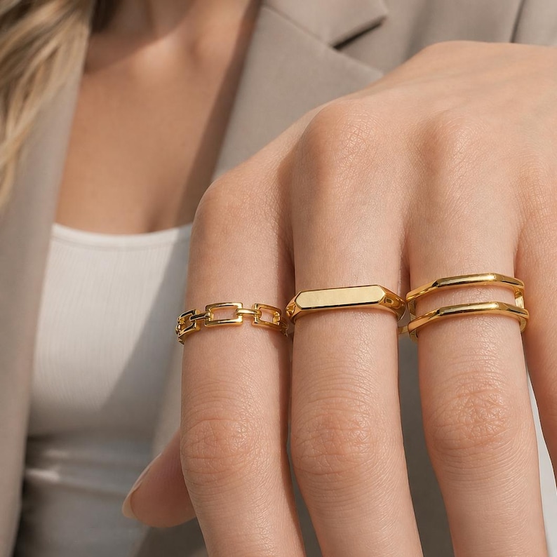 May include: Three gold-toned rings worn on fingers. One ring features a rectangular chain-link design, the second is a smooth flat-topped band, and the third is a double-layered band with a polished finish and a consistent metallic gold color.