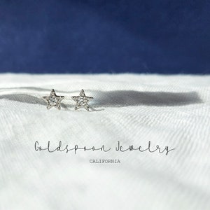 May include: A pair of silver star-shaped earrings with small clear stones set in the star shape. The earrings are on a white fabric background with the text "Goldspoon Jewelry CALIFORNIA" below the earrings.