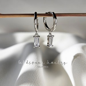 Puede incluir: Un par de pendientes de aro de plata con una piedra rectangular transparente que cuelga. Los pendientes est&aacute;n colgados de un palo de madera.