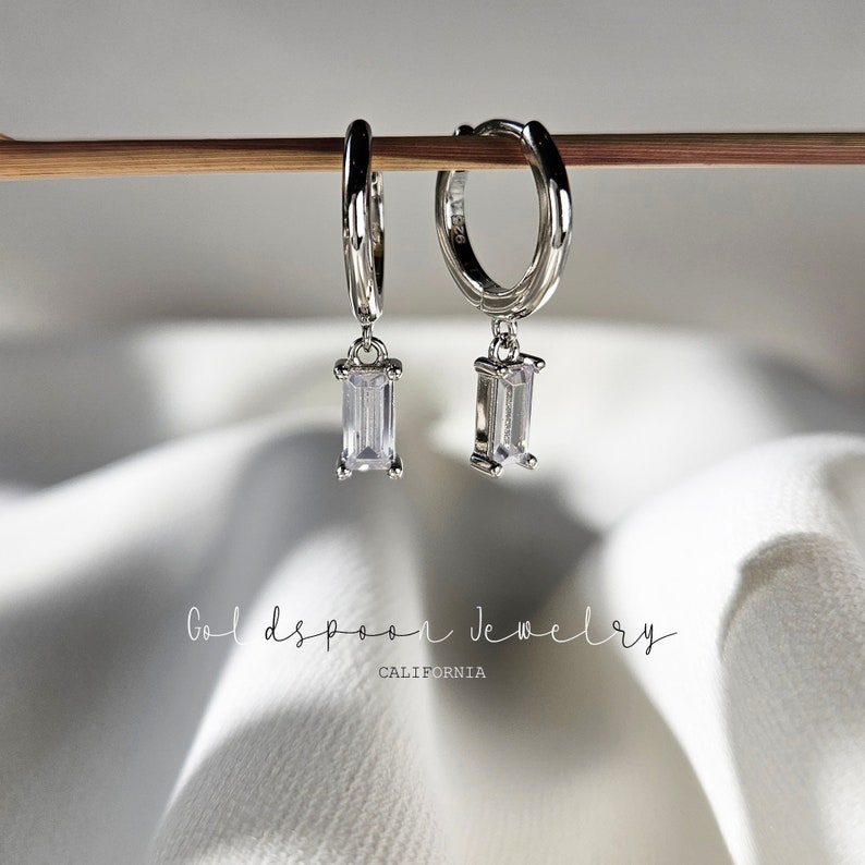 Puede incluir: Un par de pendientes de aro de plata con una piedra rectangular transparente que cuelga. Los pendientes est&aacute;n colgados de un palo de madera.