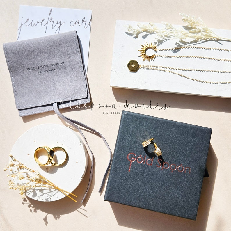 May include: Gold Spoon Jewelry packaging with a gray velvet pouch, a white stone dish with two gold rings, and a black box with gold lettering. The jewelry includes a gold necklace with a sun pendant and a hexagon pendant.