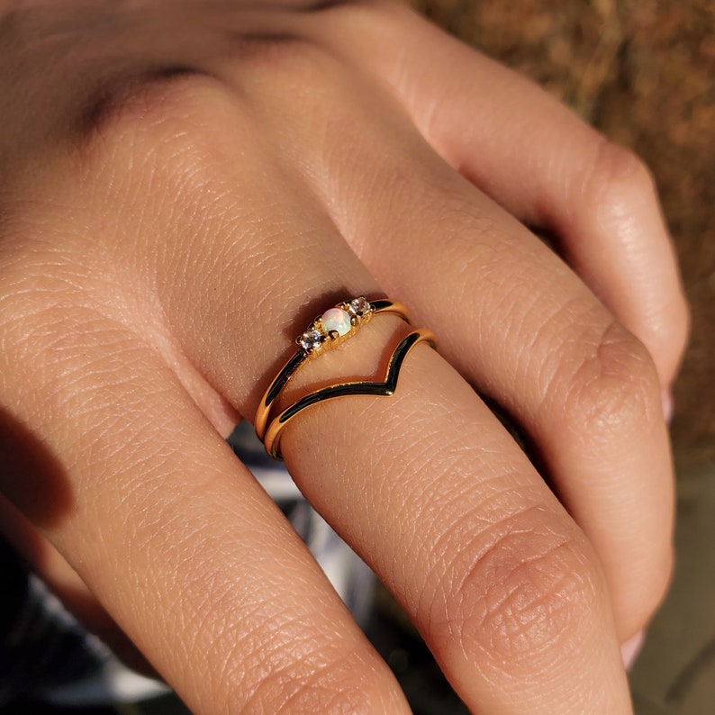 May include: A gold-colored ring with a double band design. The top band features a small opal stone flanked by two smaller clear stones. The ring is worn on a finger, showcasing its delicate details and elegant style.