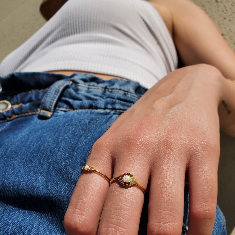 May include: Two gold rings are displayed on a hand. One ring features a small row of gemstones, while the other has a central opal surrounded by red and white stones. The rings are set against a background of blue denim.