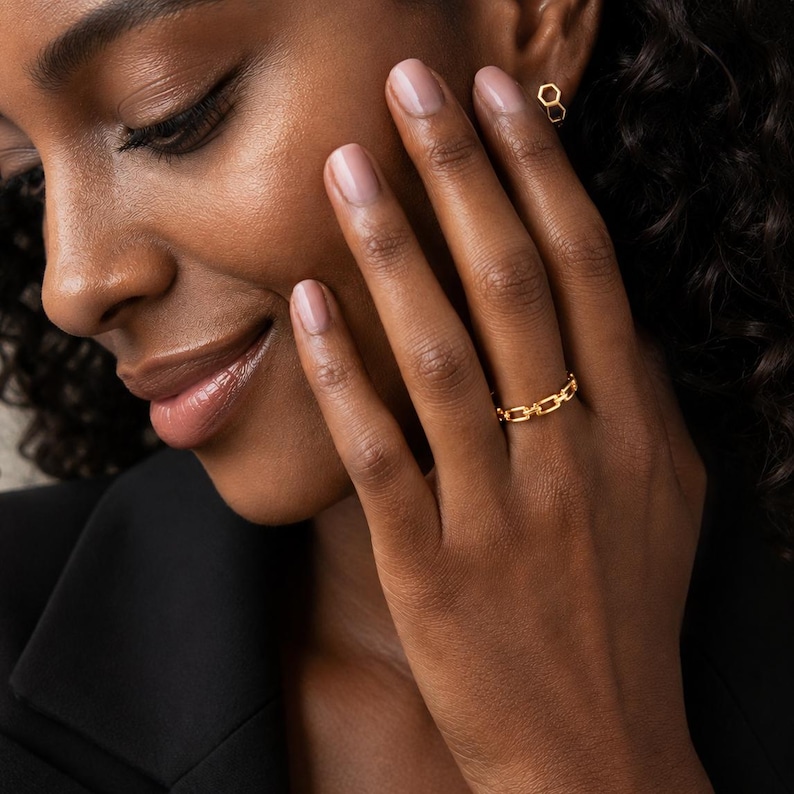 May include: A gold chain link ring featuring a series of small, interconnected rectangular loops. The ring is worn on a finger, and a small gold stud earring in the shape of a hexagon is visible on the earlobe, matching the metallic tone.