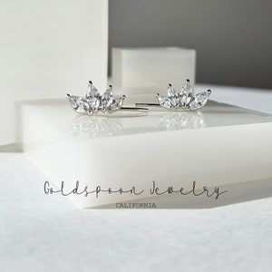 May include: A pair of silver earrings with a cluster of clear cubic zirconia stones in a crown shape. The earrings are on a white surface with the text "Goldspoon Jewelry CALIFORNIA" below.
