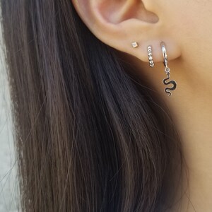 May include: A close-up of a person's ear with three earrings. The top earring is a small diamond stud. The middle earring is a small hoop with tiny beads. The bottom earring is a hoop with a snake charm hanging from it.