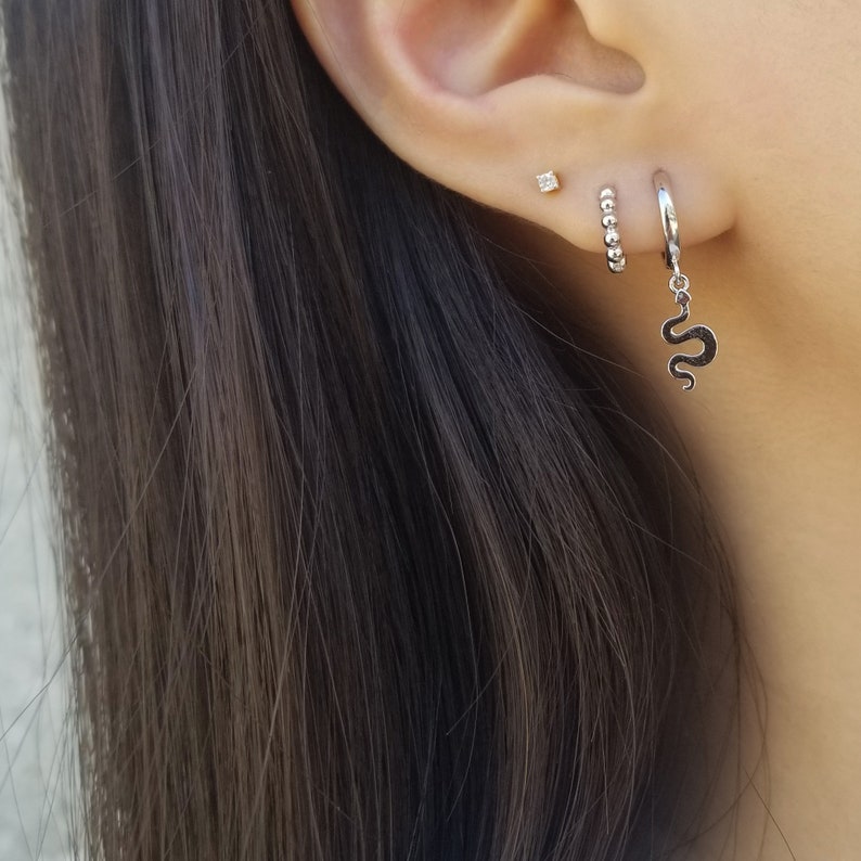 May include: A close-up of a person's ear with three earrings. The top earring is a small diamond stud. The middle earring is a small hoop with tiny beads. The bottom earring is a hoop with a snake charm hanging from it.