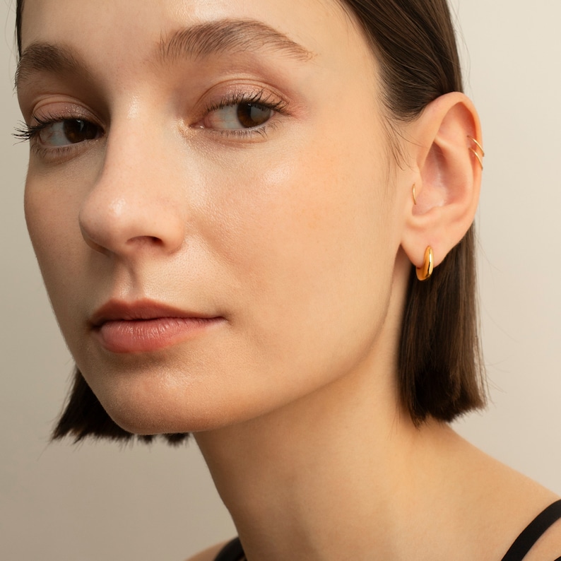 May include: A woman with short brown hair wearing gold hoop earrings in her left ear. The earrings are a simple, classic design.