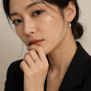May include: A silver-toned ring featuring a repeating chain-link design worn on a finger, paired with a small silver-toned hoop earring that has a single clear rectangular stone dangling from the bottom of the hoop, set against a neutral background.