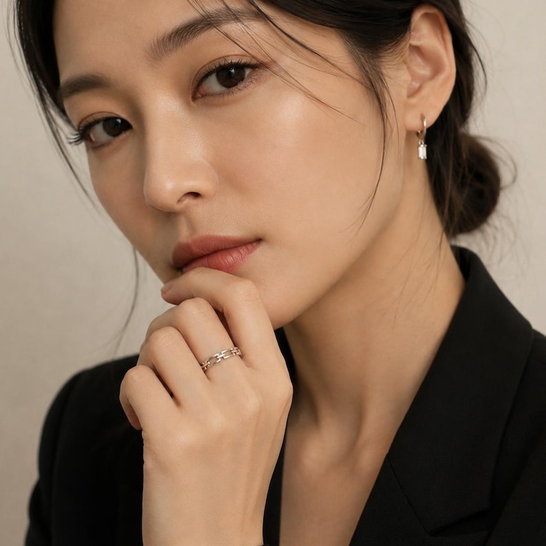May include: A silver-toned ring featuring a repeating chain-link design worn on a finger, paired with a small silver-toned hoop earring that has a single clear rectangular stone dangling from the bottom of the hoop, set against a neutral background.