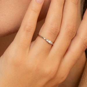 May include: A delicate silver ring featuring three small, white gemstones. The ring is thin and elegant, with the gemstones set in a row. The ring is shown on a finger, highlighting its minimalist design and subtle sparkle.