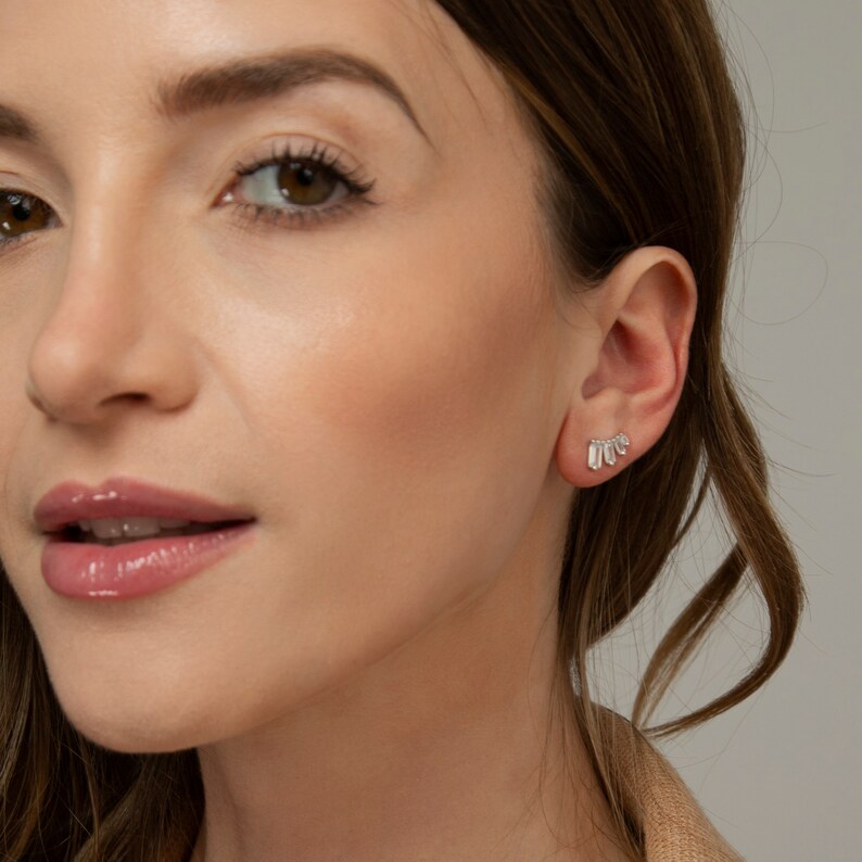 May include: A close-up of a person's face with a small, silver earring with a row of small, clear stones. The earring is in the shape of a crescent moon.