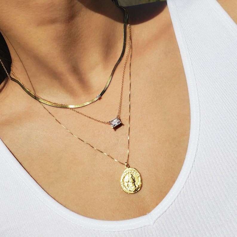 May include: Three gold necklaces layered on a person's neck. The top necklace is a thin, flat chain with a small diamond-shaped pendant. The middle necklace is a thicker chain with a larger, rectangular pendant. The bottom necklace is a chain with a round pendant featuring a detailed design.