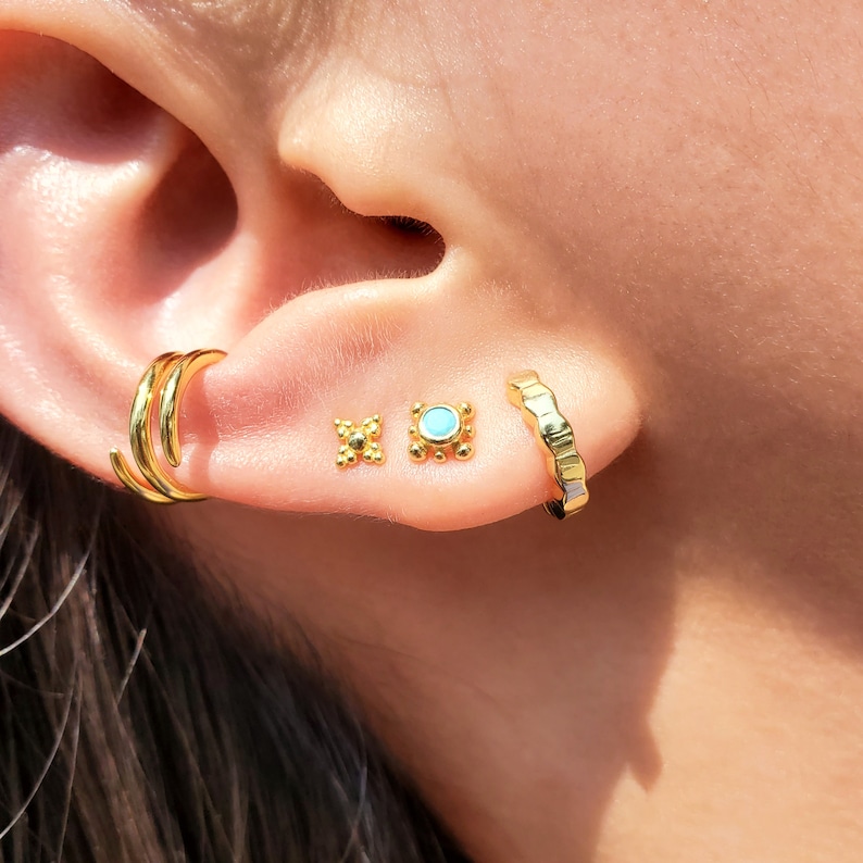 May include: A person's ear with three gold earrings. The top earring is a double hoop, the middle earring is a small square with a textured design, and the bottom earring is a small square with a turquoise stone in the center.