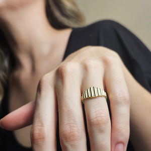 May include: A gold ring with a textured, ridged design. The ring is worn on a finger.