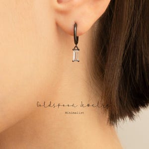 Puede incluir: Pendientes de aro negros con una piedra rectangular transparente colgante. Los pendientes son de estilo minimalista.