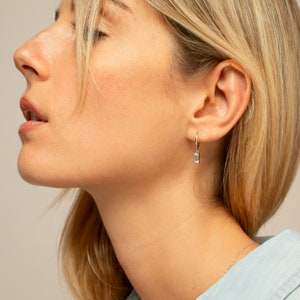 Puede incluir: Primer plano de la oreja de una mujer con un pendiente de aro de plata con una peque&ntilde;a piedra rectangular transparente que cuelga.