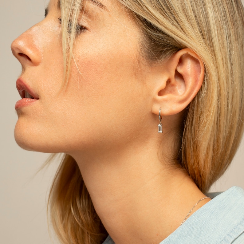 Puede incluir: Primer plano de la oreja de una mujer con un pendiente de aro de plata con una peque&ntilde;a piedra rectangular transparente que cuelga.
