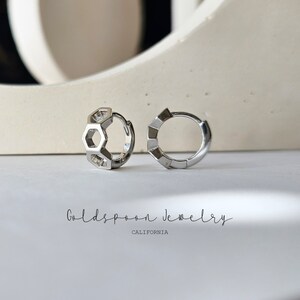 May include: A pair of silver hoop earrings. One earring features a geometric design of interlocking hexagons. The other earring is a simple hoop. The earrings are displayed on a white surface. The text "Goldspoon Jewelry California" is visible below the earrings.