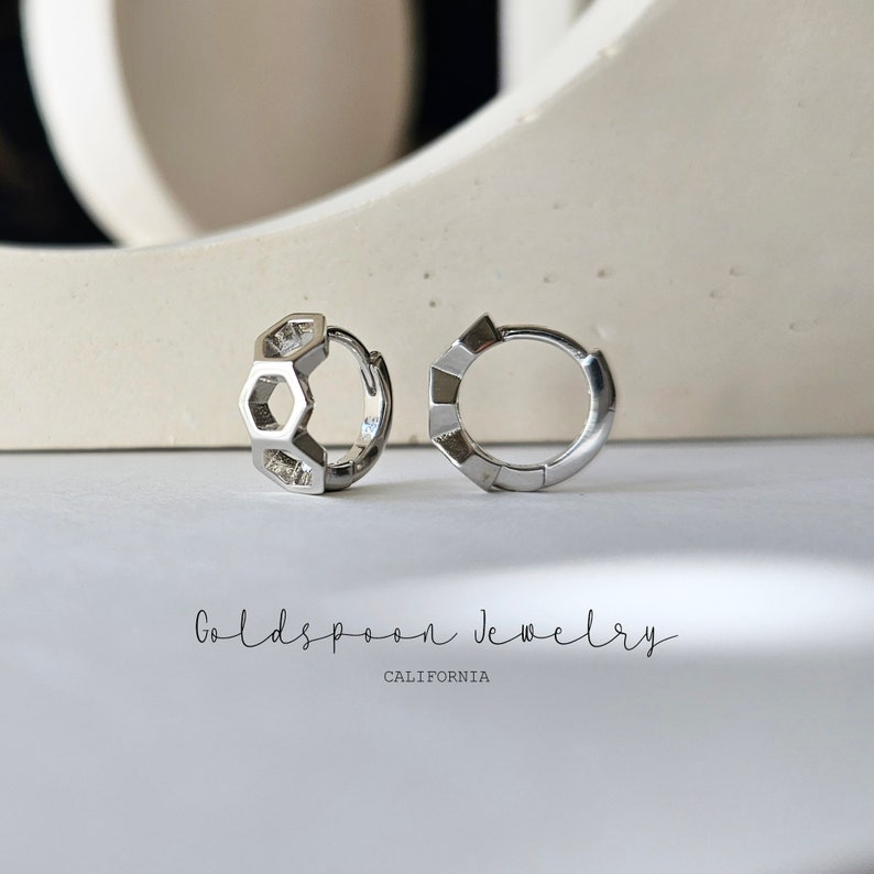May include: A pair of silver hoop earrings. One earring features a geometric design of interlocking hexagons. The other earring is a simple hoop. The earrings are displayed on a white surface. The text "Goldspoon Jewelry California" is visible below the earrings.