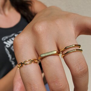 May include: Three gold rings are shown on fingers. The rings feature a chain link design, a rectangular band, and a double-banded ring. The rings have a polished finish, reflecting light. The jewellery is likely for fashion or personal adornment.