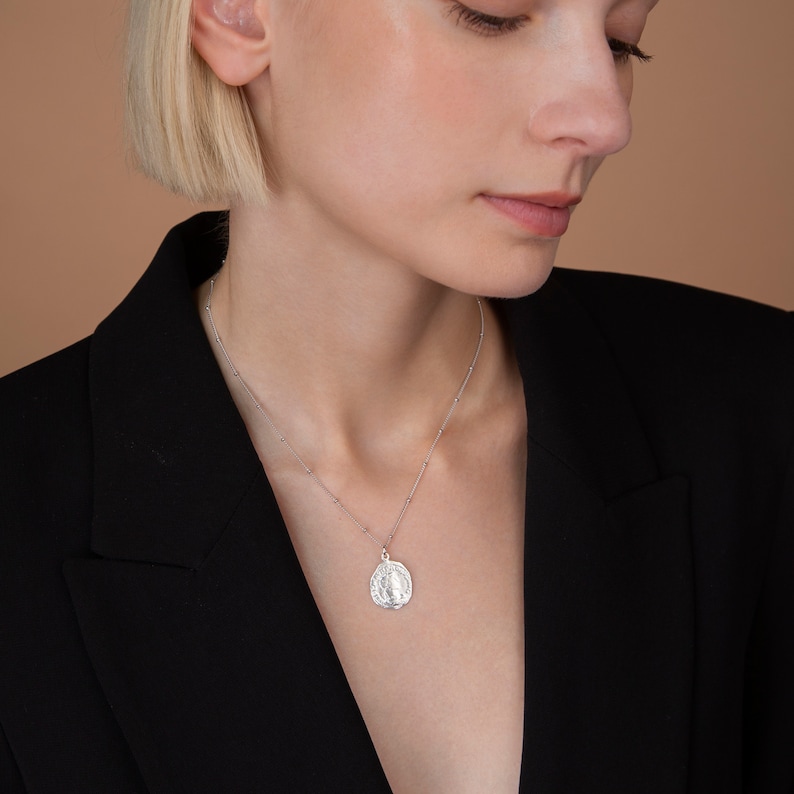 May include: A silver chain necklace with a silver pendant featuring a detailed image of the Virgin Mary. The necklace is worn by a person with short blonde hair and a black blazer.