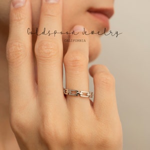 May include: A silver chain link ring. The ring is worn on a person's finger. The ring is made of a shiny metal and has a series of rectangular links. The text "Goldspoon Jewelry CALIFORNIA" is in the background.