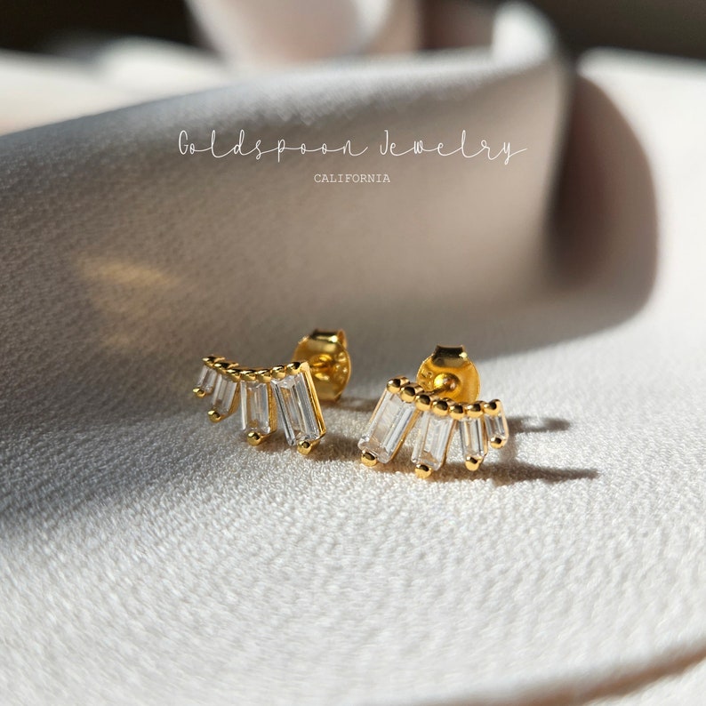 May include: Gold earrings with a row of clear baguette-shaped stones set in a curved shape. The earrings are on a white fabric background.