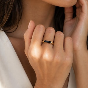 May include: A gold ring featuring a horizontal, rectangular black stone set in the center of the band. The ring has a smooth, polished finish and a clean, linear design that sits flat across the finger, showcasing a contrast between the metal.