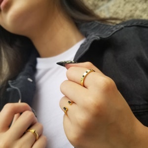 May include: A close-up of a hand wearing two gold rings. The ring on the index finger has a row of small black stones. The ring on the middle finger is a simple gold band.