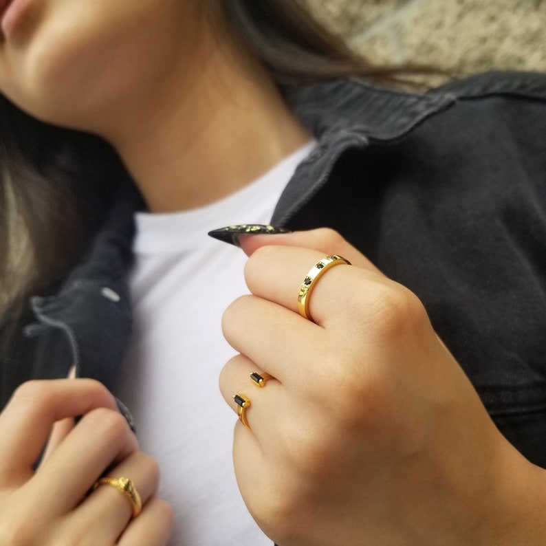 May include: A close-up of a hand wearing two gold rings. The ring on the index finger has a row of small black stones. The ring on the middle finger is a simple gold band.
