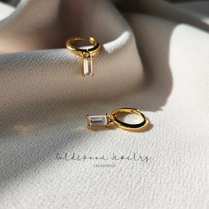 May include: A pair of gold hoop earrings with a dangling rectangular clear stone. The earrings are on a white fabric background.