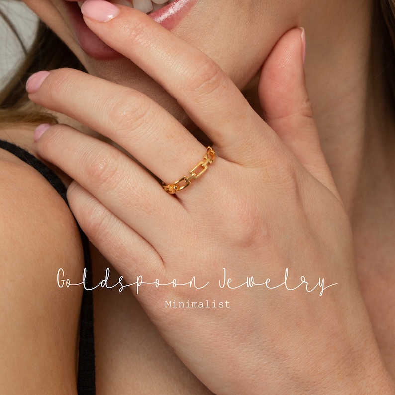May include: A gold chain link ring is worn on a finger. The ring is the focal point of the image. The text "Goldspoon Jewelry" and "Minimalist" are written in a cursive font.