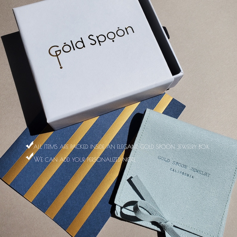 Op de afbeelding: Een lichtblauwe su&egrave;de sieradenbeurs met een gouden lint en de tekst "Gold Spoon Jewelry California" gedrukt op de voorkant. De beurs ligt op een blauw-gouden gestreept oppervlak.